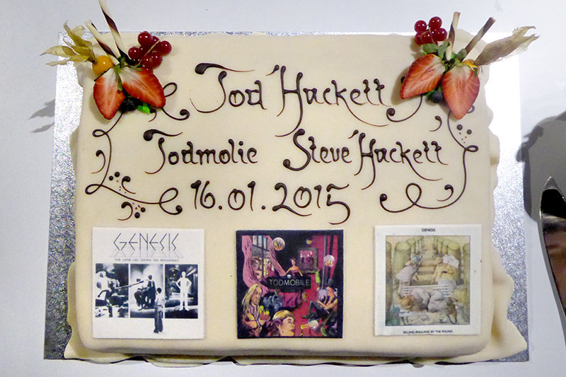 Steve Hackett Island. /></a>Am nächsten Tag ging es zum Inlandsflughafen von Reykjavik, der am Stadtrand liegt. Die Hacketts und die Band hatten schon einen früheren Flug genommen, aber es gehen täglich mehrere Flüge zwischen Reykjavik und Akureyri hin und her. Es sind kleine Propellermaschinen mit vielleicht 50 Passagieren Fassungsvermögen. Man holt sich zwar am Flughafenschalter die Bordkarte ab, aber es gibt dort keinerlei Sicherheitskontrollen. Man geht einfach durch eine Tür auf das Rollfeld und steigt in die Maschine ein. Wir hatten Glück mit dem Wetter und konnten den Flug bei blauem Himmel genießen und somit einen Blick auf endlose weiße Landschaften. Es war zwar ein Vulkan aktiv (Bárðarbunga), aber den haben wir nicht sehen können und man hätte ihn aus der Ferne wohl auch nicht von Wolken unterscheiden können, da er nur mäßig aktiv gewesen sein soll. Akureyri liegt in einem langgestreckten Fjord und es war schon kurz nach Sonnenuntergang, als wir dort landeten. Während in Reykjavik eher wenig Schnee lag, gab es hier im noch höheren Norden deutlich mehr davon und es war ca. -5 °C kalt, dafür aber nicht ganz so windig. Nach der Landung haben wir erst kurz unser Apartment bezogen, das sogar noch geräumiger war als das in Reykjavik, und sind dann zur Konzerthalle gegangen, die quasi um die Ecke lag. Die Halle, „Hof“ genannt, fasst gut 500 Zuschauer, ist aber topmodern und gut ausgestattet. Nachdem wir dort im Foyer erst mal mit den Hacketts gespeist hatten, sind wir direkt mit ihnen in den Saal gegangen, wo es noch einen letzten Soundcheck gab. Für diese Show hatte ich einen Platz in der 2. Reihe ergattert, aber ganz außen. Es gab aber auch weiter hinten unterhalb des Oberrangs ein paar freie Plätze, mehr in der Mitte, wo ich mich dann niederließ und den noch besseren Sound (im Vergleich zum Vortag) genießen konnte. Die Setliste war genau die gleiche, auch die Besetzung, nur dass sich Chor und Orchester teilweise aus anderen Musikern von Akureyri zusammensetzten.</p>
<p> <a title=